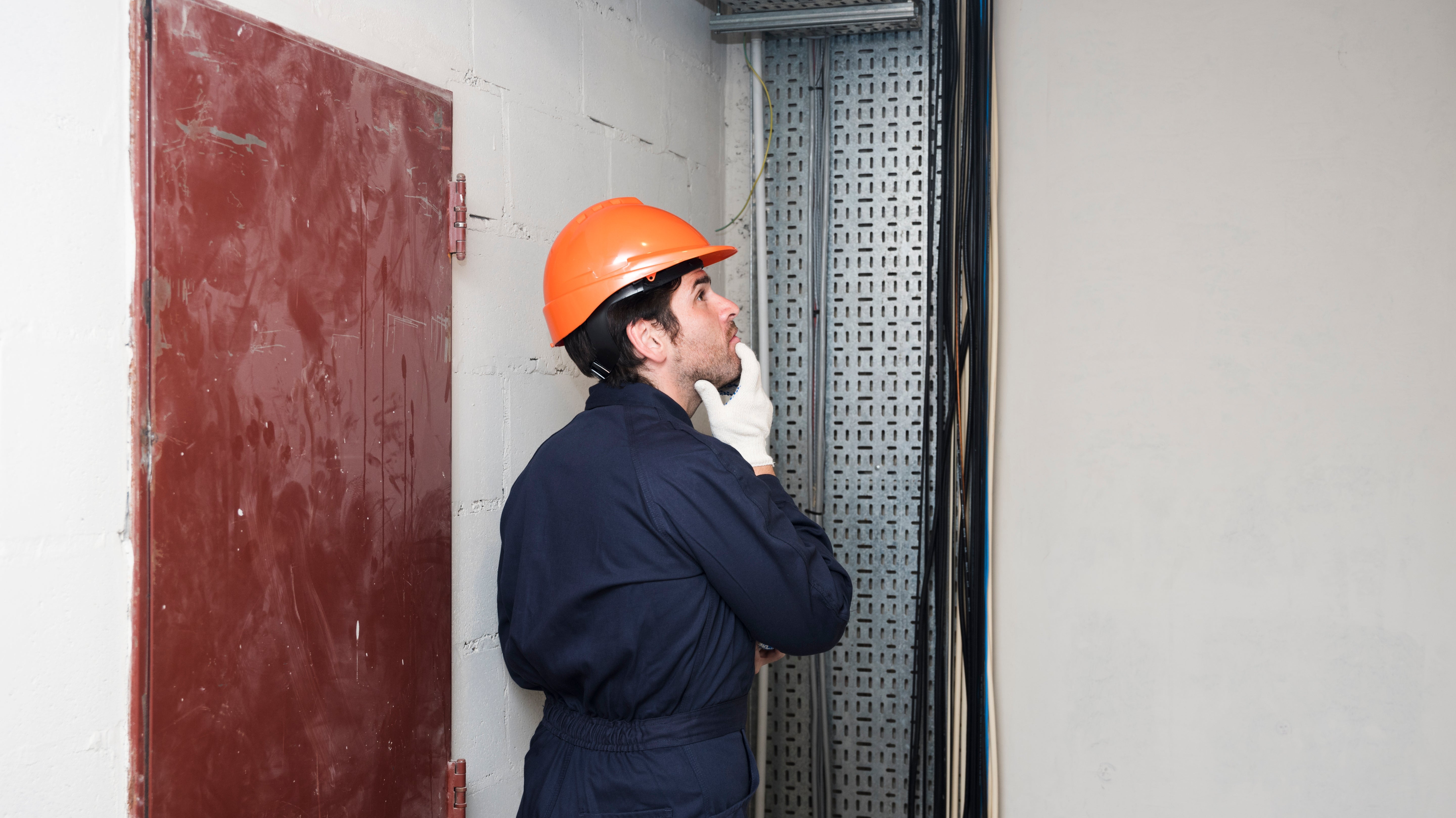 Electrician in orange hard hat inspecting fire door and wiring in industrial setting