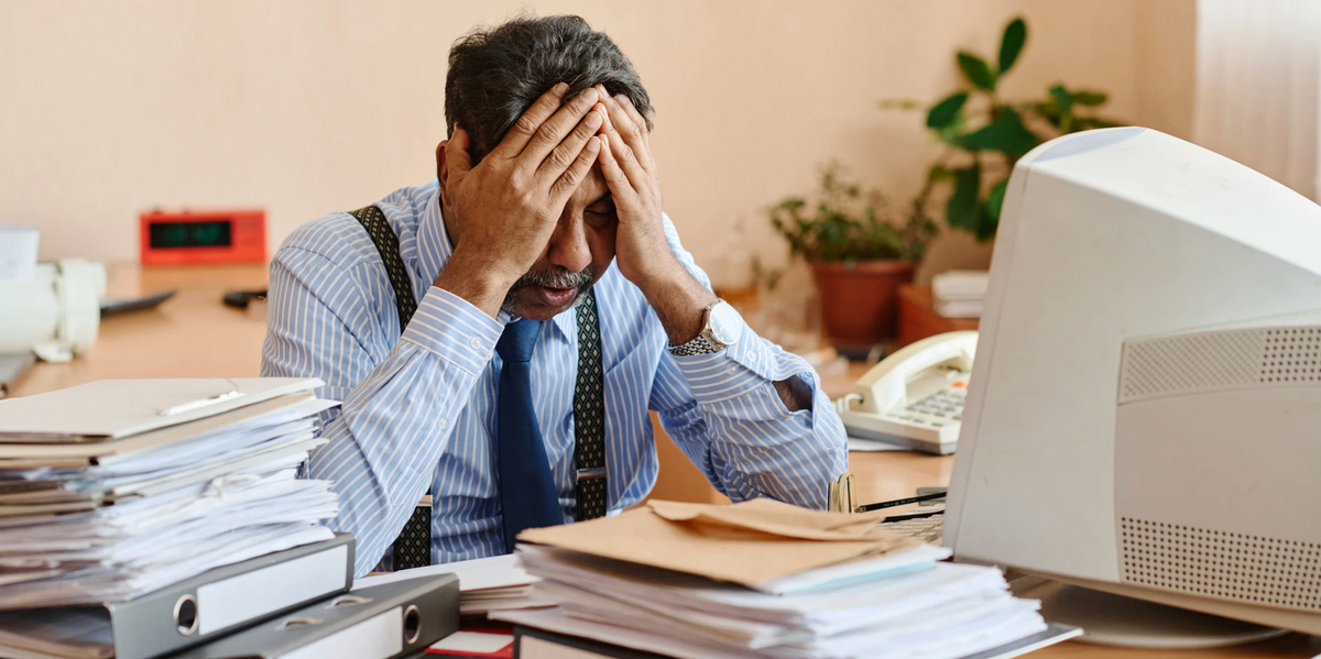 Stressed office worker with head in hands at cluttered desk with documents and computer